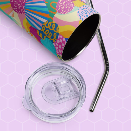 The Squiggs and Stripes Stainless Steel Tumbler from My Favourite Colour is Rainbow, featuring a vibrant abstract pattern, lies on its side with a clear lid and sleek metal straw nearby. The background shows a pastel pink honeycomb pattern.