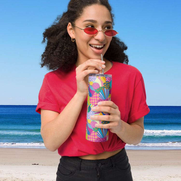 A woman in a red shirt and small red sunglasses smiles on a sandy beach under a clear sky, holding the colorful Squiggs and Stripes Stainless Steel Tumbler with a metal straw by My Favourite Colour is Rainbow, with the calm blue ocean behind her.