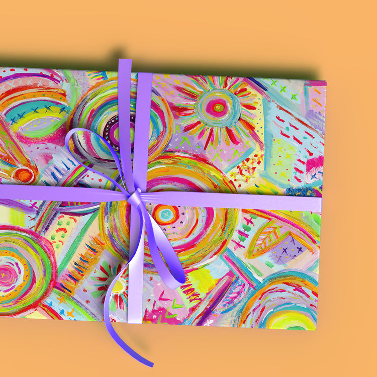 A gift box wrapped in Summer Sorbet Wrapping Paper Sheets from My Favourite Colour is Rainbow, featuring bright abstract circular patterns in pink, yellow, green, and blue. It is tied with a shiny purple ribbon on an orange background.
