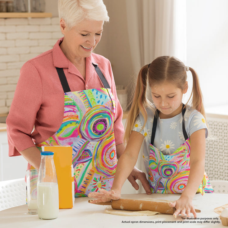 In the kitchen, a woman and a young girl bake together in colourful Summer Sorbet Aprons by My Favourite Colour is Rainbow. The girls is rolling dough, with a milk bottle and juice carton close by. The apron features abstract colourful circles, lines, and geometric patterns in neon hues.