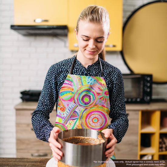 A person with blonde hair wears a Summer Sorbet Apron by My Favourite Colour is Rainbow, as they prepare dessert in a modern kitchen. They hold a cake tin, looking down at their baking amidst yellow cabinets and sleek appliances, showcasing culinary creativity.  The apron features abstract colourful circles, lines, and geometric patterns in neon hues.