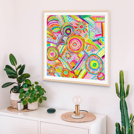 Summer Sorbet Fine Art Print by My Favourite Colour is Rainbow, with circular patterns and a certificate of authenticity, hangs above a sideboard with a small plant, decorative bulb, and smart speaker. A tall cactus stands beside the furniture.