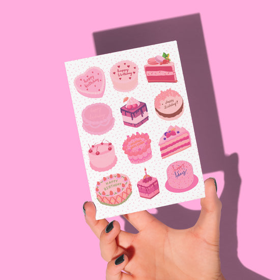 A hand with dark nail polish holds the Sweet Treats Birthday Card by My Store, featuring cake designs with pink treats like slices and whole cakes. Each cake displays Happy Birthday messages on a soft pink background.