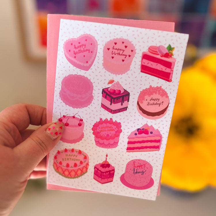 A hand holds My Stores Sweet Treats Birthday Card with charming cake designs, cherries, and strawberries, displaying Happy Birthday messages. A colorful painting and a yellow flower add an artistic touch in the background.