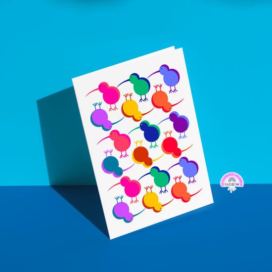 The Technicolour Kiwi greeting card from My Store displays stylized kiwi birds in pink, blue, green, orange, and purple on a white background. Its perfect for brightening someone’s day as it casts a shadow on a split blue surface.