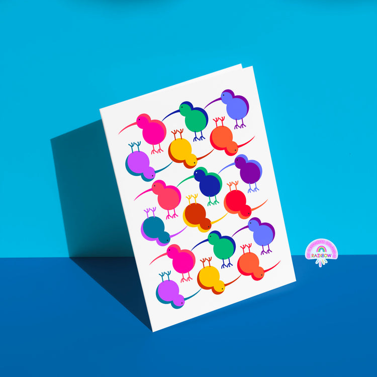 The Technicolour Kiwi greeting card from My Store displays stylized kiwi birds in pink, blue, green, orange, and purple on a white background. Its perfect for brightening someone’s day as it casts a shadow on a split blue surface.