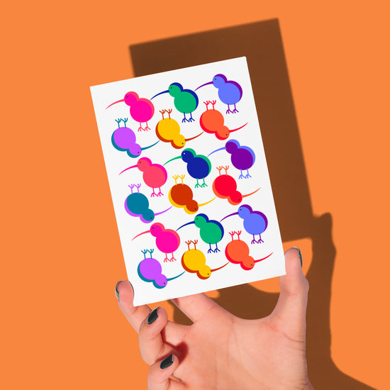 A hand with painted nails holds a Technicolour Kiwi greeting card by My Store, featuring vibrantly colored abstract birds on a white background, set against a solid orange backdrop.