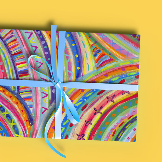 A gift clad in The Rainbow Connection Wrapping Paper from My Favourite Colour is Rainbow, featuring abstract rainbow designs and a light blue ribbon. Shown on a yellow background.