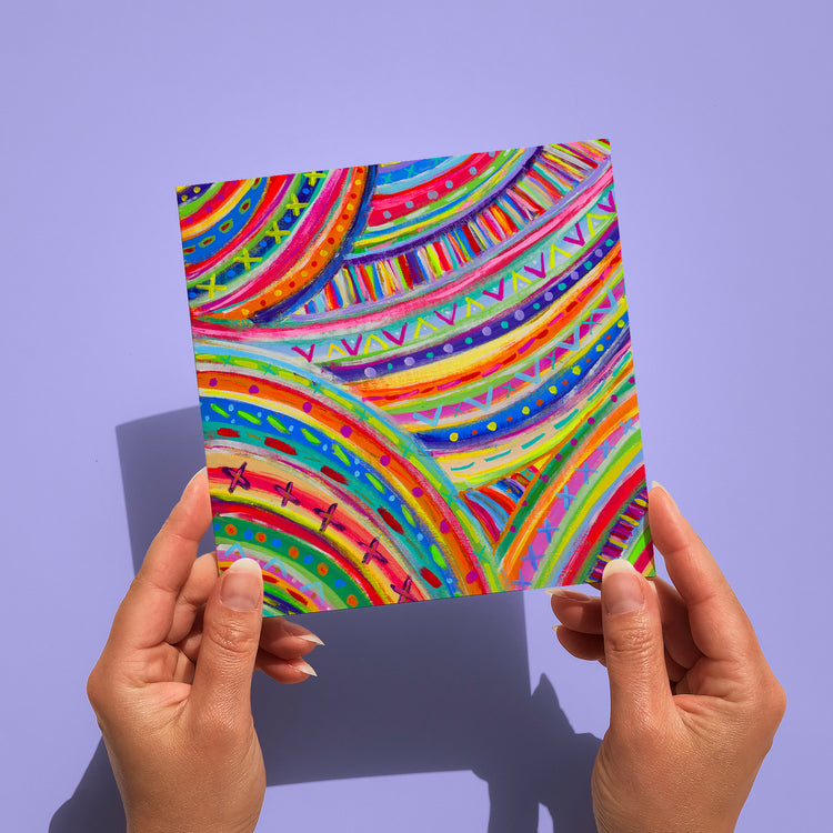 Two hands hold The Rainbow Connection Greeting Card by My Store against a light purple background. This artwork, printed on high-quality card stock, features colorful curved patterns with stripes, dots, and zigzags in vibrant pink, yellow, blue, and green.
