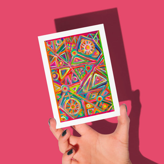 A hand with black nails holds My Stores Tropical Geometry Greeting Card against a pink backdrop, featuring bold triangles, circles, and vivid colors such as pink, green, and orange.