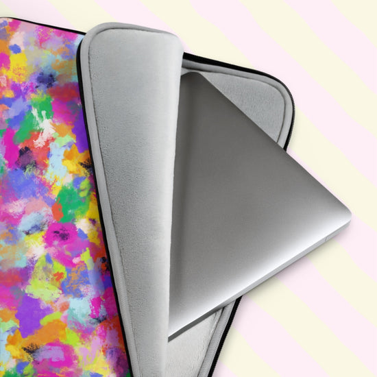 A close-up of a silver laptop partially out of the Watercolour Rainbow Laptop Sleeve by My Favourite Colour is Rainbow, showcasing its vibrant abstract pattern with pink, purple, blue, yellow, and green splashes of colour.  It has a grey padded interior to keep your laptop safe from scratches.  The print is on only one side of the laptop sleeve, the other side is black. It is shown on a pastel background.