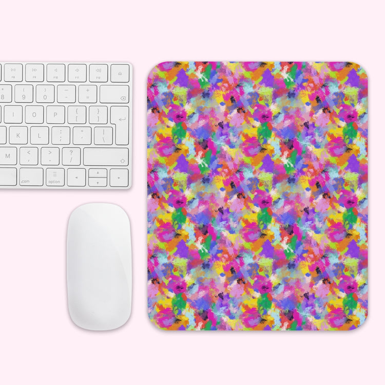 The Watercolour Rainbow Mouse Pad by My Favourite Colour is Rainbow features a vibrant abstract paint splatter design. It’s shown on a light pink background with a white wireless mouse and keyboard.