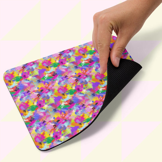 A hand lifts the corner of a Watercolour Rainbow Mouse Pad by My Favourite Colour is Rainbow, featuring a vibrant abstract watercolor design on top and a black rubber underside, placed against a light background.