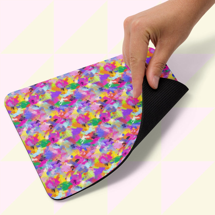 A hand lifts the corner of a Watercolour Rainbow Mouse Pad by My Favourite Colour is Rainbow, featuring a vibrant abstract watercolor design on top and a black rubber underside, placed against a light background.