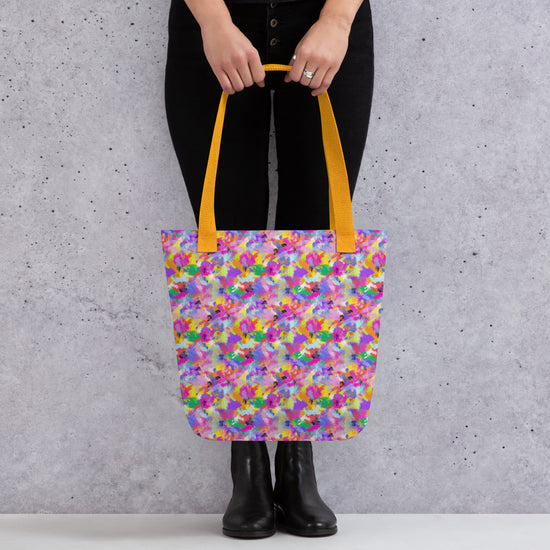A person holds the Watercolour Rainbow Tote Bag by My Favourite Colour is Rainbow, showcasing its vibrant abstract design in purple, yellow, and green with dual orange handles. They wear black pants and boots against a gray wall.
