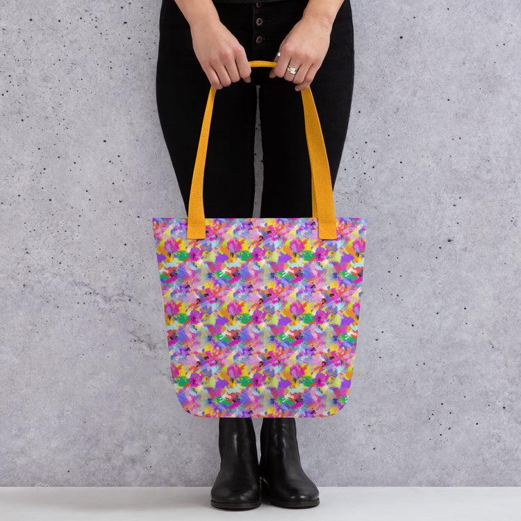 A person holds the Watercolour Rainbow Tote Bag by My Favourite Colour is Rainbow, showcasing its vibrant abstract design in purple, yellow, and green with dual orange handles. They wear black pants and boots against a gray wall.