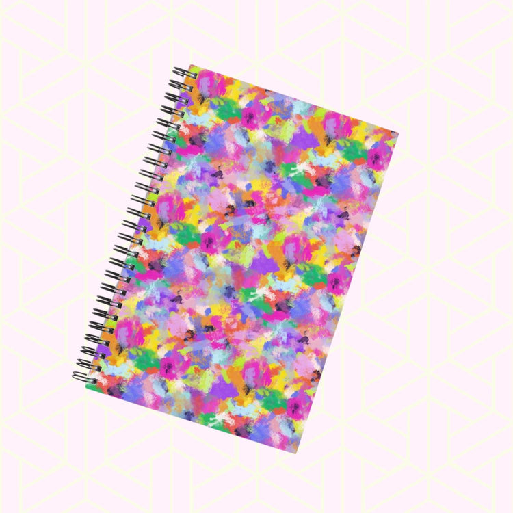 The Watercolour Rainbow Notebook by My Store features a spiral-bound design with a vibrant paint-splatter cover in pink, yellow, green, blue, and purple. Positioned diagonally on a light geometric patterned backdrop, its essential for any creative stationery collection.