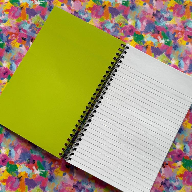 A Watercolour Rainbow Notebook by My Store sits open against a vibrant, abstract background. The left page is solid green while the right is lined. The lively backdrop of pink, yellow, purple, and blue splashes ignites creativity.