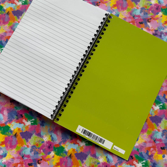 A Watercolour Rainbow Notebook by My Store sits open on an abstract background; its left page has horizontal lines while the right cover is solid green with a barcode sticker at the bottom, inviting creativity.