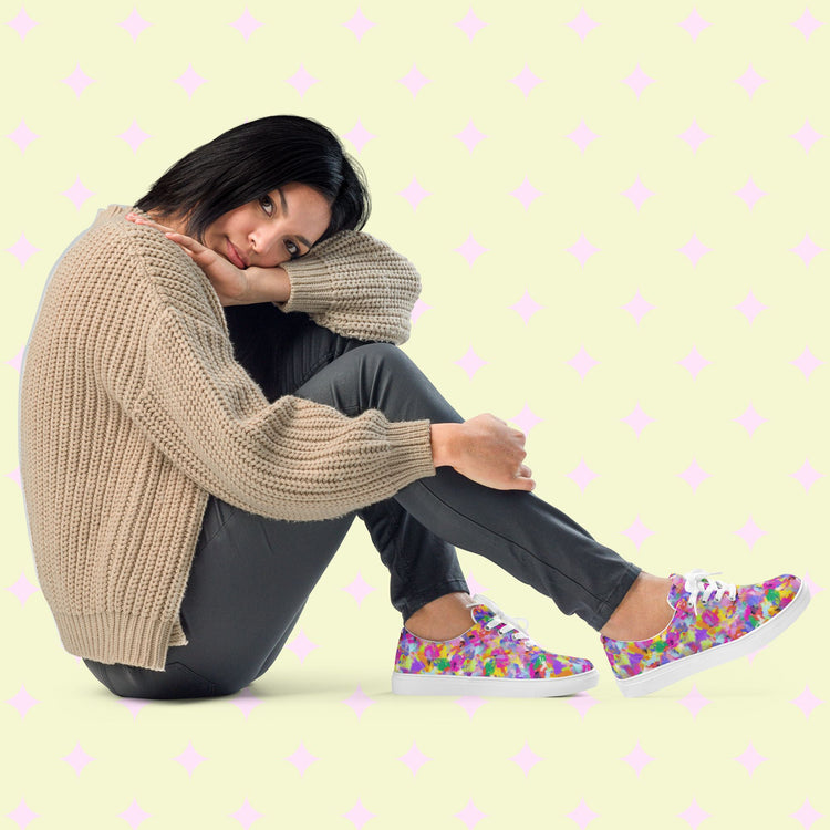 A person with short black hair sits on the floor, head resting on their knee. They wear a beige sweater, gray pants, and Watercolour Rainbow Canvas Sneakers by My Favourite Colour is Rainbow. The light green background has pink diamond shapes and a watercolor rainbow effect.