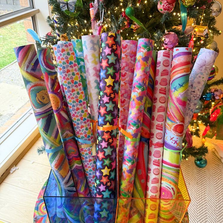 Displayed in a stand are rolled wrapping paper sheets by My Favourite Colour is Rainbow, featuring colourful abstract art and designs like stars, hearts, and stripes. A Christmas tree is in the background.