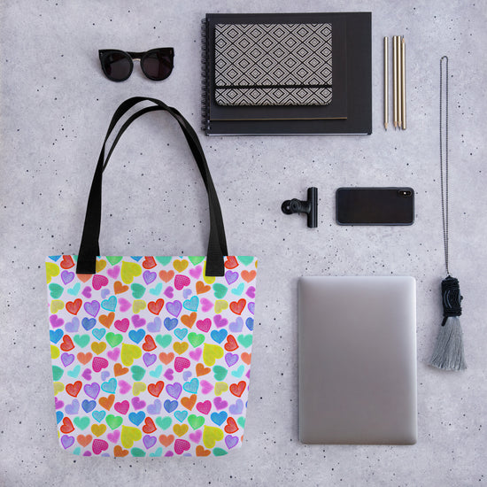 Flat lay on a speckled surface: a Heart Attack Tote Bag by My Favourite Colour is Rainbow, sunglasses, notebooks, pens, a smartphone, a laptop, a tassel accessory, and a clip-on smartphone lens.