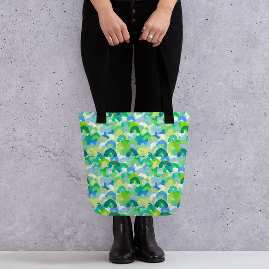 A person stands against a grey wall, holding My Favourite Colour is Rainbow;s Green Rainbow Tote Bag with a colourful green, blue, and teal pattern. They wear black pants and boots, effortlessly showcasing the bags spacious style.