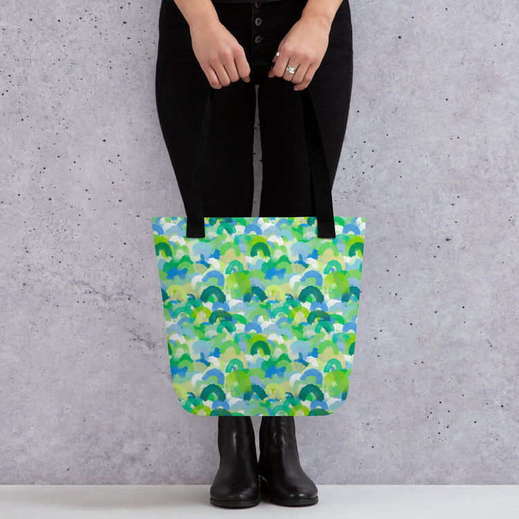 A person stands against a grey wall, holding My Favourite Colour is Rainbow;s Green Rainbow Tote Bag with a colourful green, blue, and teal pattern. They wear black pants and boots, effortlessly showcasing the bags spacious style.