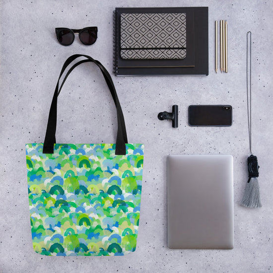 Flat lay of assorted items on a grey background: a Green Rainbow Tote Bag by My Favourite Colour is Rainbow, sunglasses, a patterned notebook, closed laptop, smartphone, clip, pens, and tassel keychain.