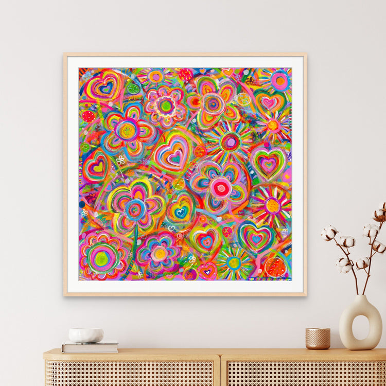 The Love in Neon Fine Art Print by My Store showcases vibrant heart and flower shapes, framed on a white wall. Available as fine art prints with a certificate of authenticity, it complements a wicker console adorned with a vase of cotton stems.