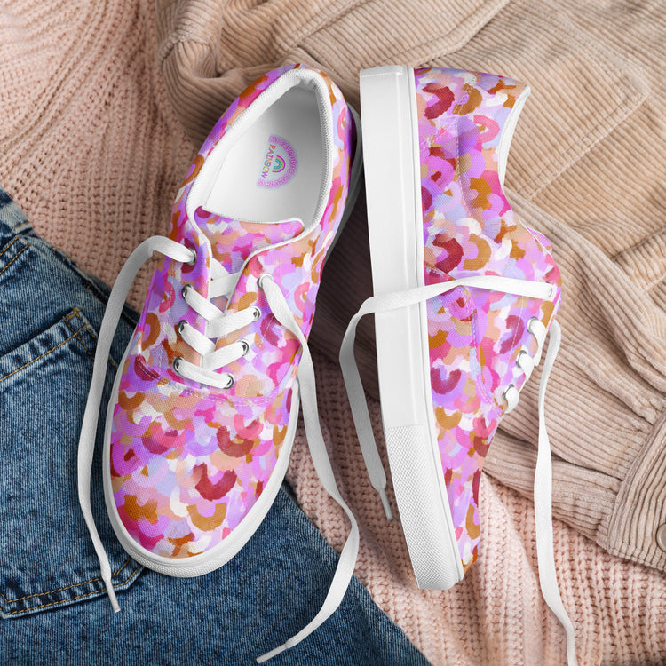 The Pink Rainbow Canvas Sneakers by My Favourite Colour is Rainbow rest on a textured beige blanket. One shoe stands upright, the other leans on its side, showcasing their vibrant style. A hint of jeans peeks from the corner.