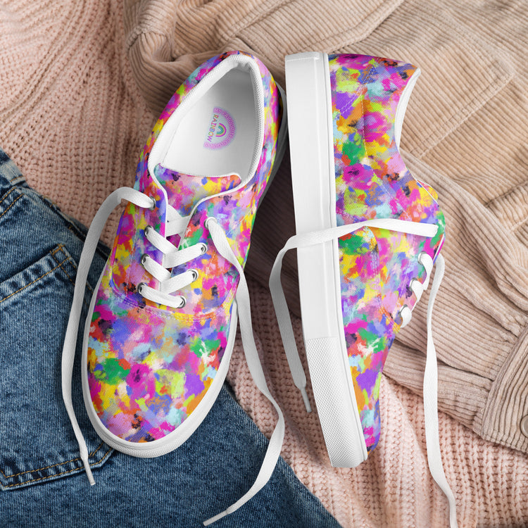 Watercolour Rainbow Canvas Sneakers by My Favourite Colour is Rainbow, featuring a purple, pink, yellow, and green pattern, rest on a knitted beige sweater. One sneaker stands upright while the other lies on its side with part of blue jeans in view.