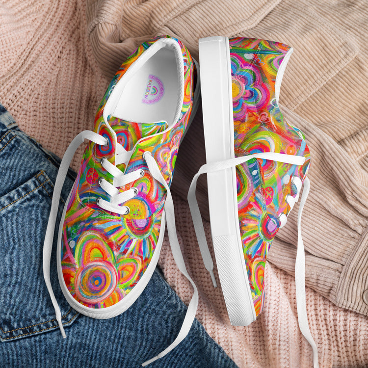 The Love in Neon Canvas Sneakers by My Favourite Colour is Rainbow showcase intricate, vibrant prints of circles and shapes. With white laces and soles, theyre set against soft pink fabric beside blue denim, creating a casual artistic setting with a vibrant aesthetic.