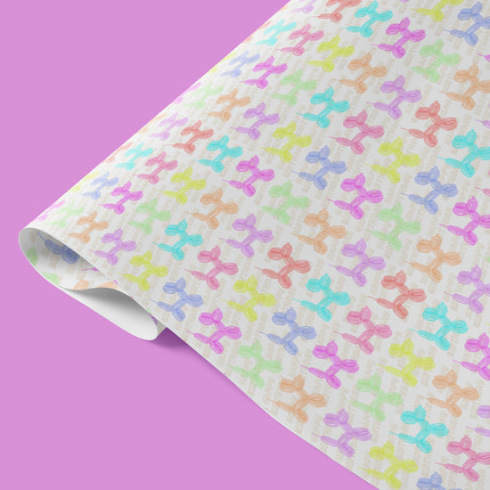 The Balloon Dog Birthday Wrapping Paper Sheets by My Favourite Colour is Rainbow feature colourful, pastel balloon animal patterns on a light background with "Happy Birthday " written in balloons diagonally, behind the balloon animals. The wrapping paper is partially rolled and is shown against a solid lavender background.