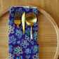 A Bloomscape Napkin by My Favourite Colour is Rainbow, with a blue floral pattern, cradles a gold fork and spoon on a round wooden plate for an elegant dining experience.