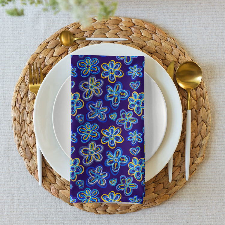 A dining setup features a white plate on a woven placemat with the Bloomscape Napkin by My Favourite Colour is Rainbow, featuring a design of large flowers and small hearts in shades of blue and yellow on a royal blue background, atop the plate. Gold cutlery flanks each side.