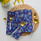 The Bloomscape Set of 4 Napkins from My Favourite Colour is Rainbow, featuring yellow and blue florals, adorns a round woven placemat. Gold-hued utensils, like forks and spoons, are arranged around the napkins for an elegant dining touch. Yellow flowers add charm to the scenes top.