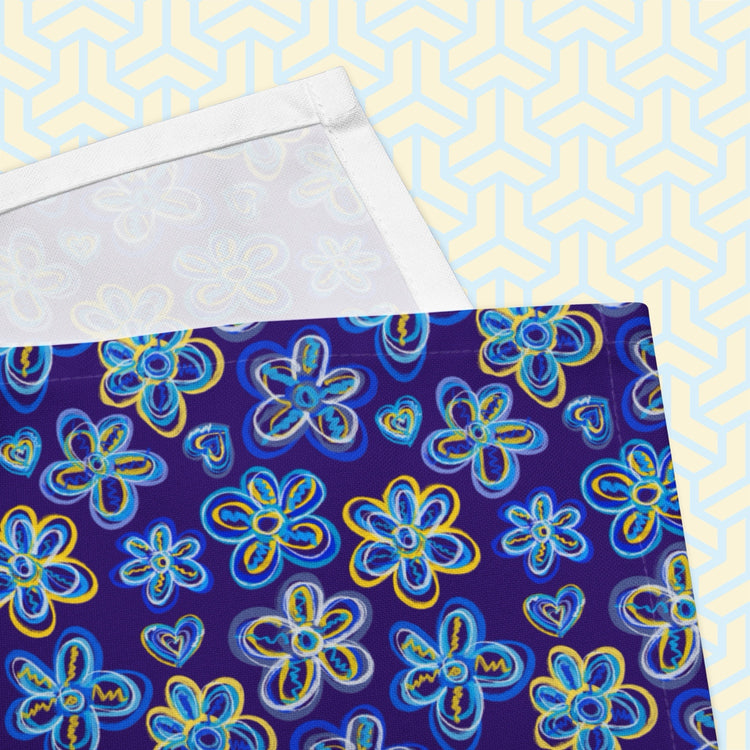 A close-up highlights the vibrant charm of My Favourite Colour is Rainbows Bloomscape Napkins. The napkin features a design of large flowers and small hearts in shades of blue and yellow on a royal blue background.  The print is on only one side of the napkin; the other side is plain white.