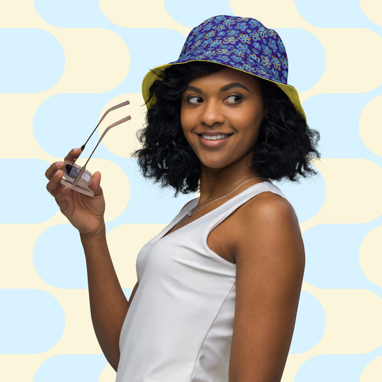 A woman in a white tank top holds sunglasses, wearing the Bloomscape Reversible Bucket Hat by My Favourite Colour is Rainbow.  The hat features a design of large flowers and small hearts in shades of blue and yellow on a royal blue background.