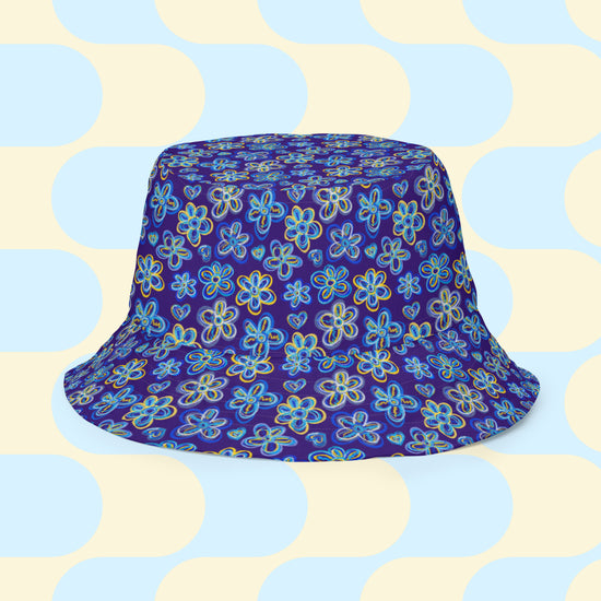 The Bloomscape Reversible Bucket Hat by My Favourite Colour is Rainbow features a design of large flowers and small hearts in shades of blue and yellow on a royal blue background. Made from breathable fabric, it combines style and comfort.