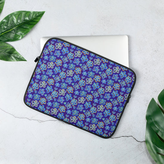 A laptop peeks out from the 13 inch Bloomscape Laptop Sleeve by My Favourite Colour is Rainbow, featuring a design of large flowers and small hearts in shades of blue and yellow on a royal blue background. It lies on a textured light surface, encircled by green leaves for a natural vibe.