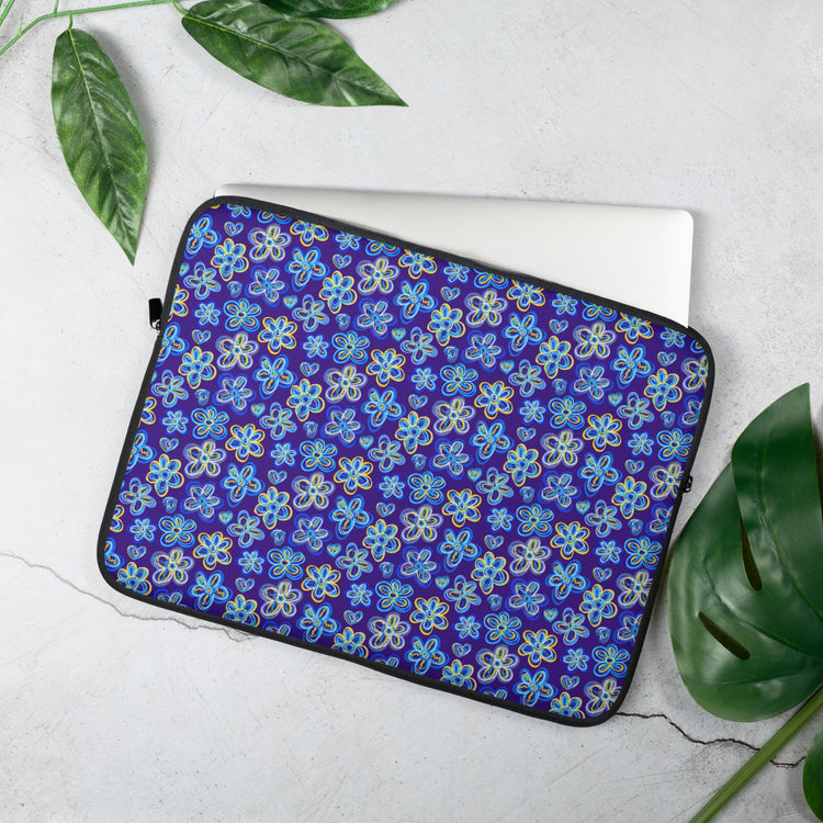 A laptop peeks out from the 15 inch Bloomscape Laptop Sleeve by My Favourite Colour is Rainbow, featuring a design of large flowers and small hearts in shades of blue and yellow on a royal blue background. It lies on a textured light surface, encircled by green leaves for a natural vibe.