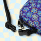 A close-up of a Bloomscape Mini Crossbody Bag from My Favourite Colour is Rainbow, showcasing a vibrant floral-inspired purple design. It features black zippers, adjustable straps with plastic buckles, and is water-resistant. A light blue and beige checkered pattern is in the background.