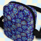 The Bloomscape Mini Crossbody Bag by My Favourite Colour is Rainbow features a vibrant floral pattern with blue and yellow flowers on dark purple, water-resistant fabric. It has adjustable black straps, a zippered closure, and a light blue and cream checkered backdrop.