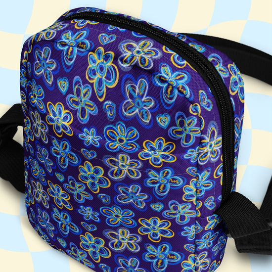 The Bloomscape Mini Crossbody Bag by My Favourite Colour is Rainbow features a vibrant floral pattern with blue and yellow flowers on dark purple, water-resistant fabric. It has adjustable black straps, a zippered closure, and a light blue and cream checkered backdrop.