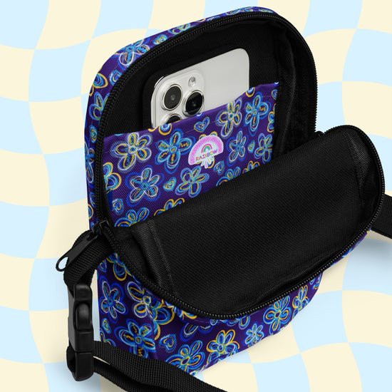 The Bloomscape Mini Crossbody Bag by My Favourite Colour is Rainbow, showcasing a vibrant floral-inspired print, opens to reveal a smartphone in its front pocket, against a striking blue and yellow checkered background.