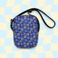 The Bloomscape Mini Crossbody bag by My Favourite Colour is Rainbow is a small rectangular accessory with a vibrant blue and yellow floral pattern on dark purple, made from water-resistant fabric. It has a black strap and an adjustable light blue and cream checkered backdrop.