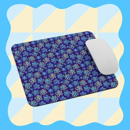 A white computer mouse sits on the Bloomscape Mouse Pad by My Favourite Colour is Rainbow, featuring a colorful floral pattern and a checkered yellow and blue background with wavy edges.