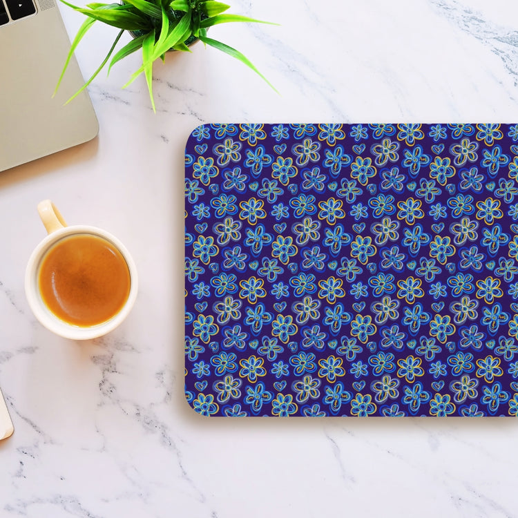 A laptop is on a marble surface beside a cup of coffee and a My Favourite Colour is Rainbow Bloomscape Mouse Pad, featuring blue and yellow floral patterns on dark. A plant is partially visible in the top left corner.