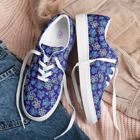 Bloomscape Canvas Sneakers by My Favourite Colour is Rainbow, featuring a blue floral pattern, breathable lining, and white soles, rest on a bed. They are paired with a light pink knitted sweater and blue denim jeans, creating a casual and cozy setting.